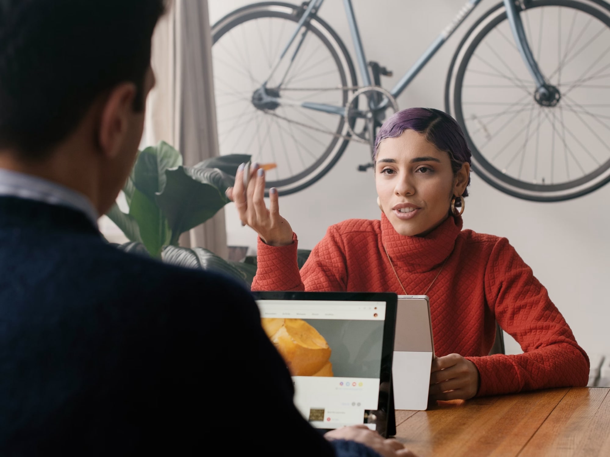 Mujer con suéter rojo hablando con un hombre mientras usa una tableta en una mesa de madera, con una bicicleta colgada en la pared de fondo.